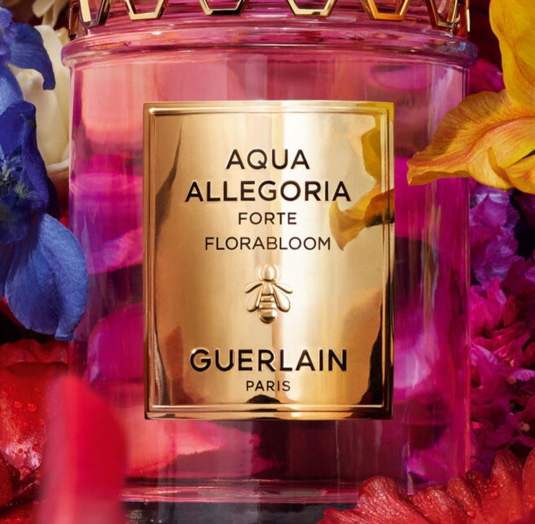 A close-up of a Guerlain Aqua Allegoria Forte Florabloom candle in a pink glass bottle with a shiny gold label, surrounded by colorful flowers.