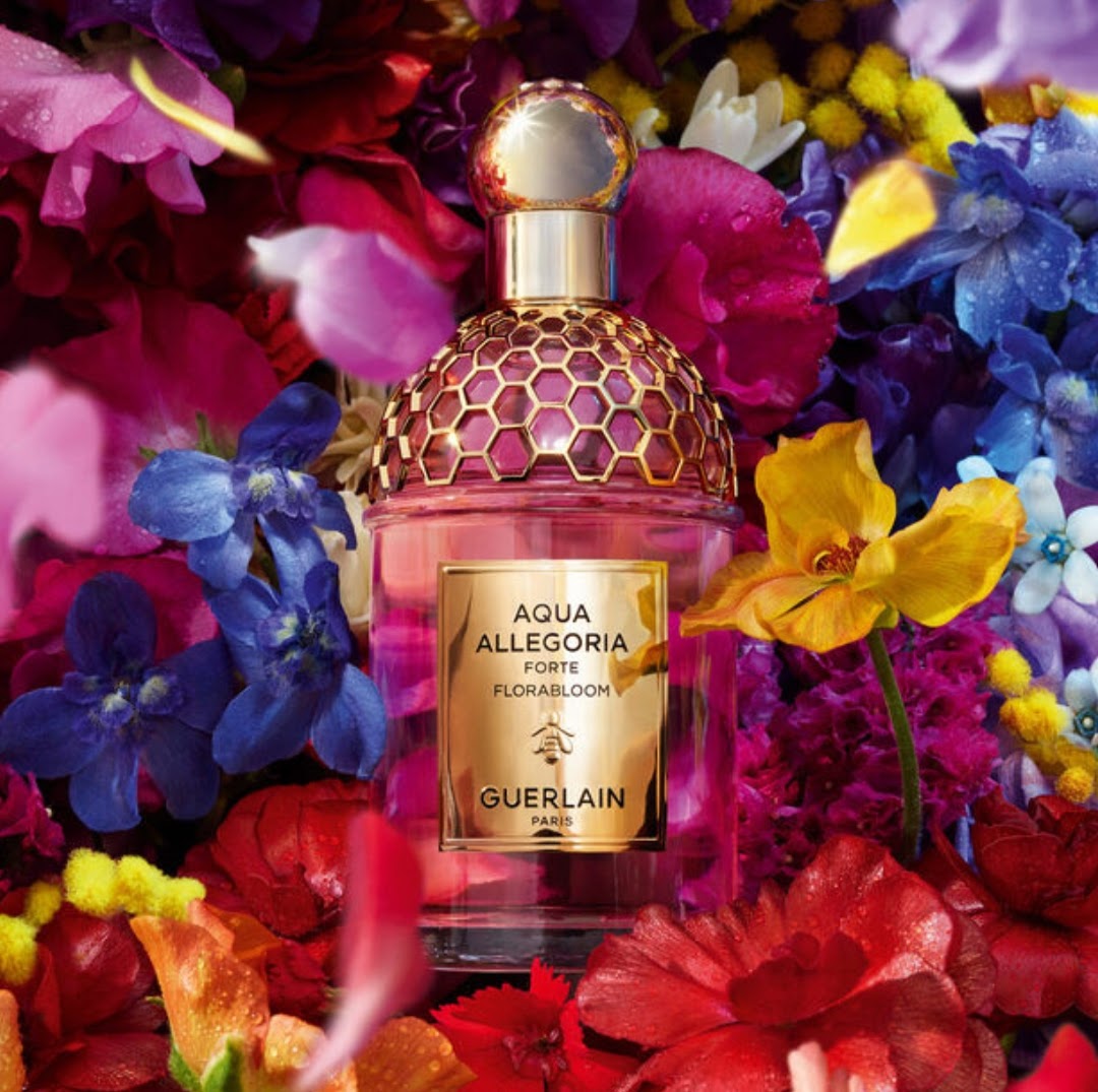Guerlain Aqua Allegoria Forte Florabloom perfume bottle surrounded by a dense bed of colorful flowers, including red, pink, blue, purple, and yellow blossoms.