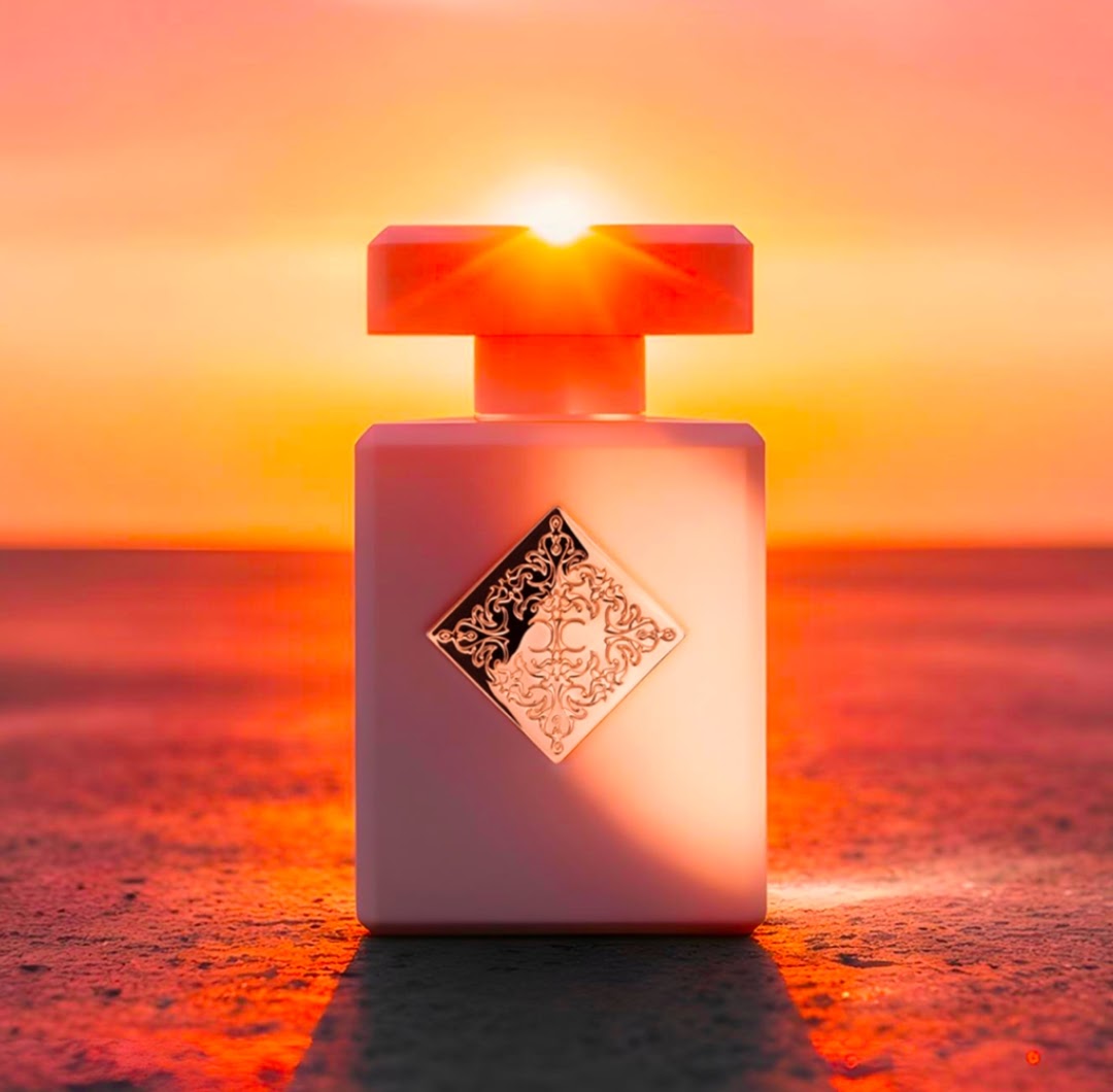 A Initio Lift Me Up perfume bottle with a square pale pink body and a matching cap, featuring an ornate gold diamond-shaped emblem on the front. The bottle is photographed outdoors at sunset with a bright orange sky and warm light reflecting off its surface.