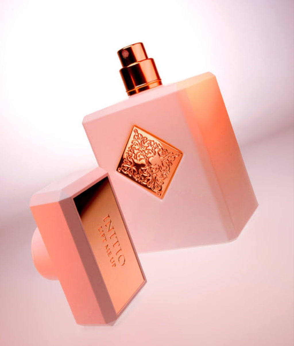 A pink rectangular Initio Lift Me Up perfume bottle with a metallic rose-gold spray top and a decorative embossed label, shown alongside its matching pink cap on a soft pink background.