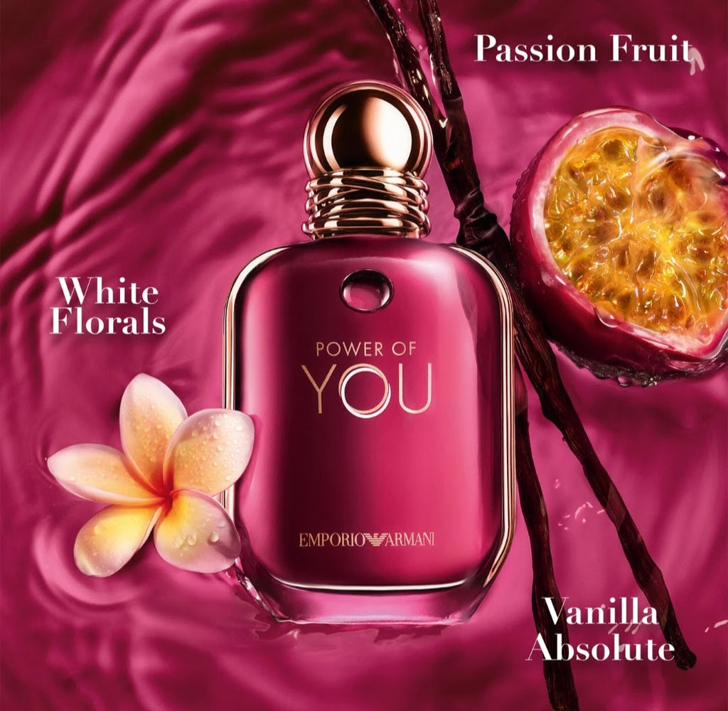 Emporio Armani Power of You Vanilla Absolute fragrance bottle in a rich magenta-pink glass with gold-toned detailing, set against a matching silky magenta background with a passion fruit half, a white frangipani flower, and text labels 'Passion Fruit', 'White Florals', and 'Vanilla Absolute'.