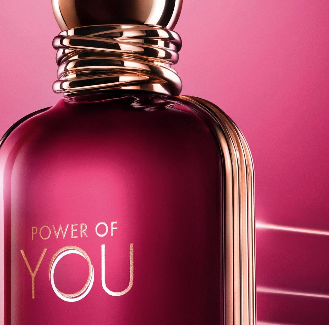 A close-up of a glossy magenta-pink perfume bottle with metallic rose-gold detailing and the text 'POWER OF YOU' on the front, set against a matching pink background.