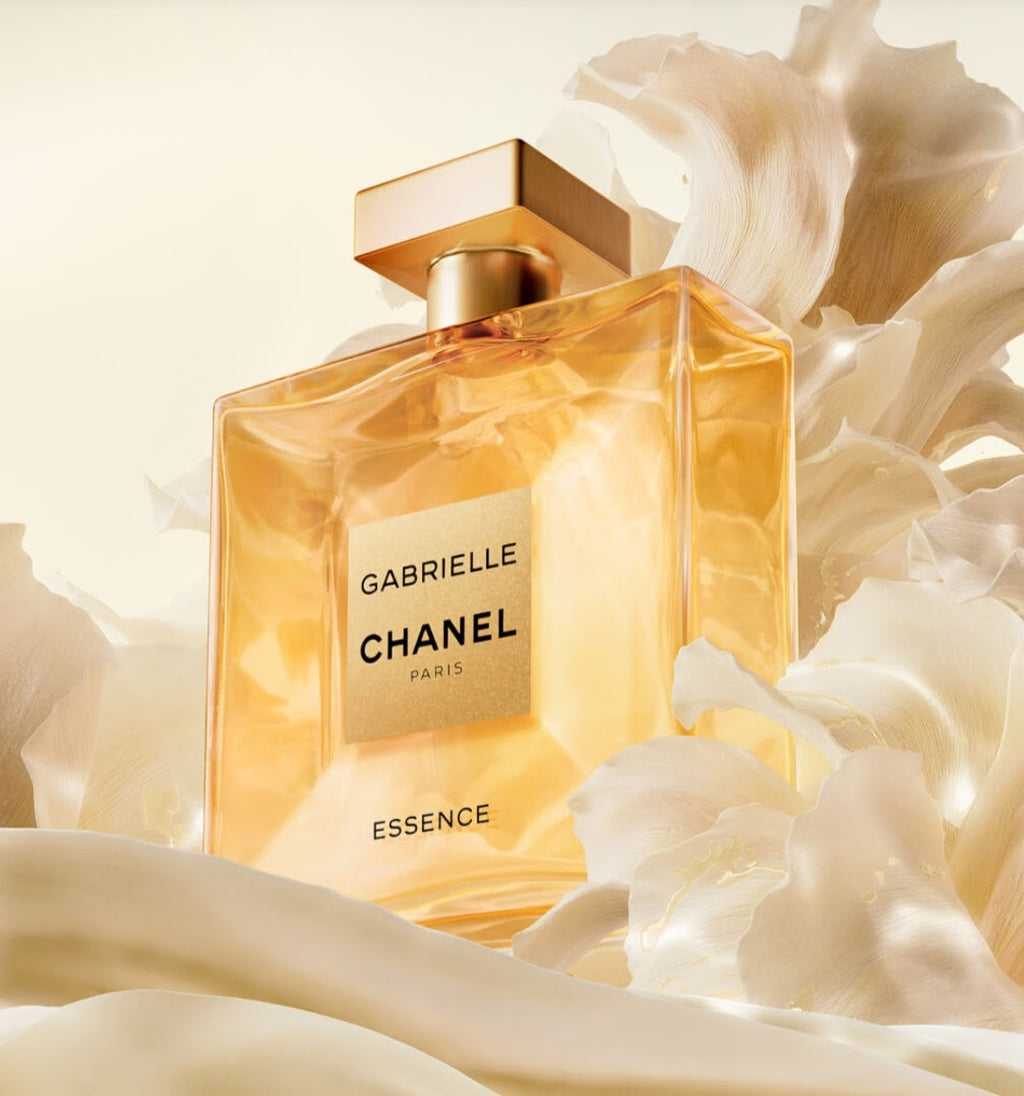 Chanel Gabrielle Essence perfume bottle in a warm amber glass with a gold cap, surrounded by soft white floral petals on a light cream background.