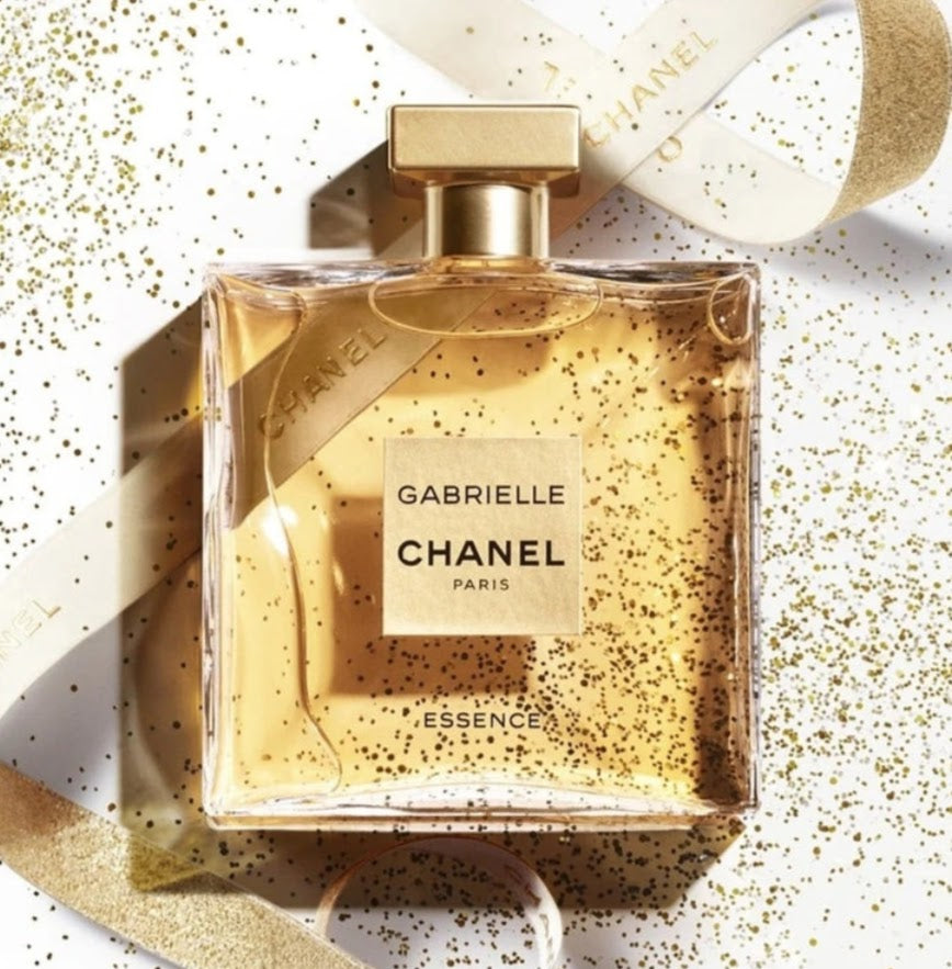 Chanel Gabrielle Essences perfume bottle with a gold cap, filled with golden liquid and decorated with gold glitter, shown with a white ribbon on a white background with gold glitter.