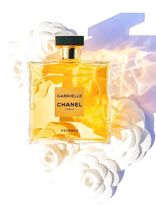 Chanel Gabrielle Essence perfume bottle with a black cap and gold label, set against a white background with stylized rose illustrations and a purple-blue graphic shape.