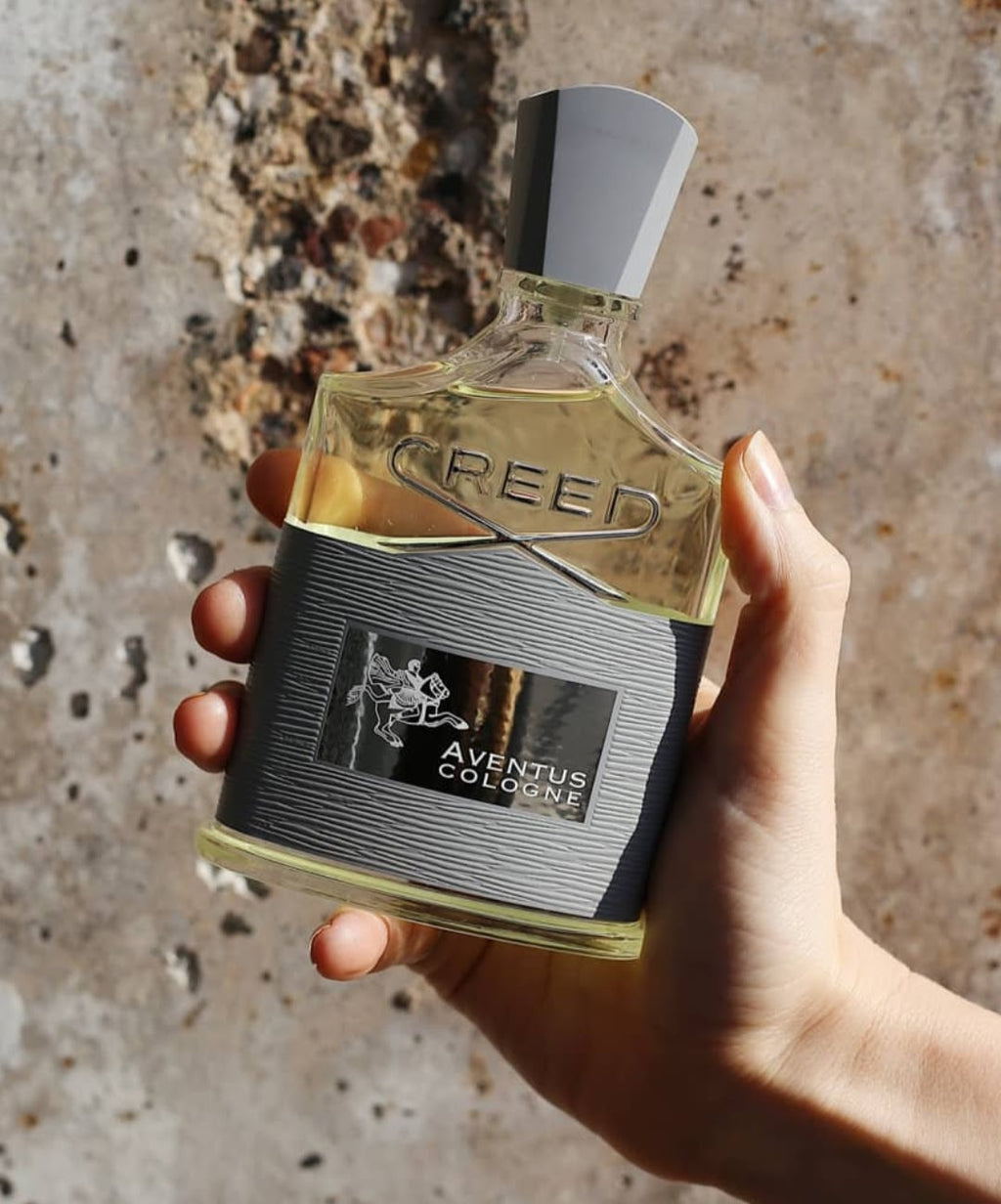A hand holding a bottle of Creed Aventus Cologne against a textured stone background.