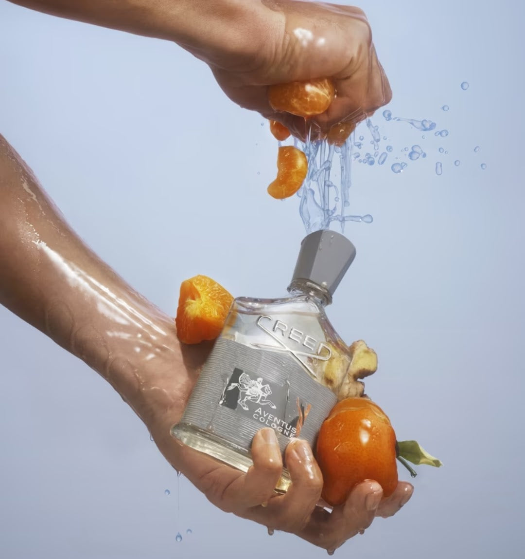 A clear glass perfume bottle labeled Creed Aventus is held underwater while being squeezed over with orange segments, causing water droplets to splash. The background is a solid light blue-gray.