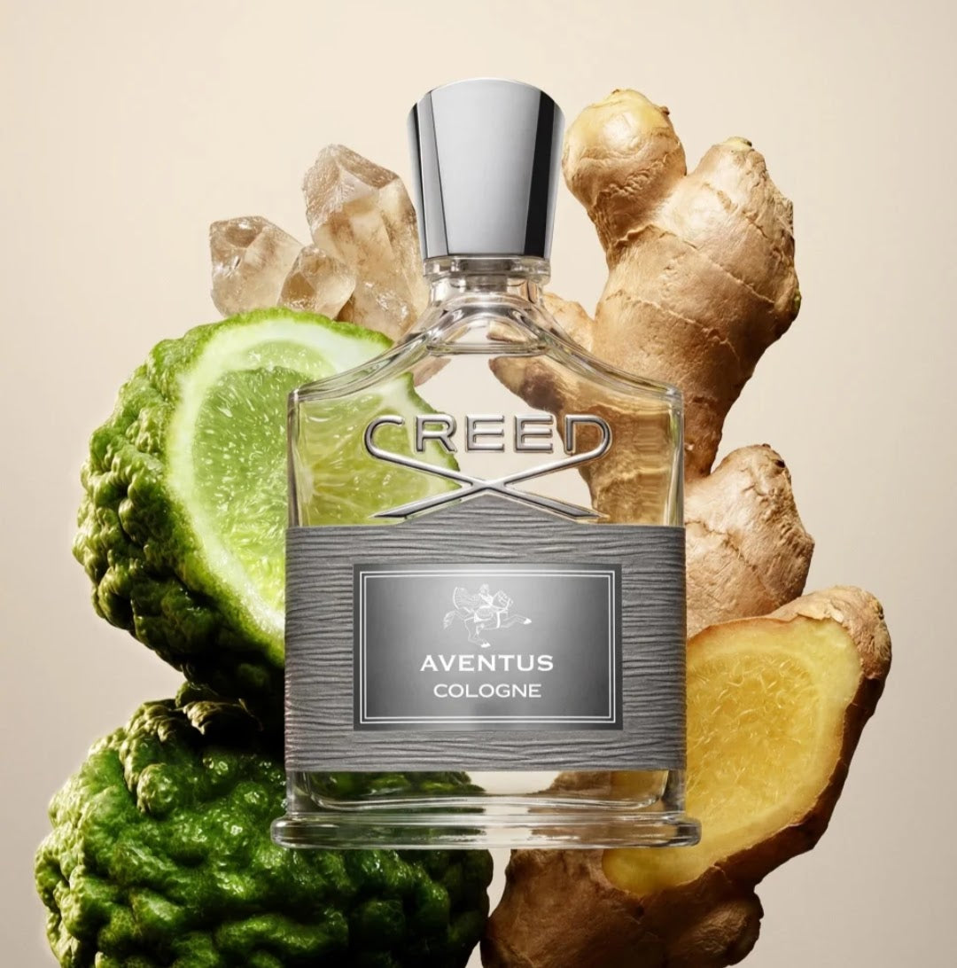 A bottle of Creed Aventus Cologne centered against a warm beige background, surrounded by fresh ingredients including ginger roots, a lime slice, and a bitter melon. The bottle is clear glass with a silver cap and a textured silver label reading 'CREED' and 'AVENTUS COLOGNE'.