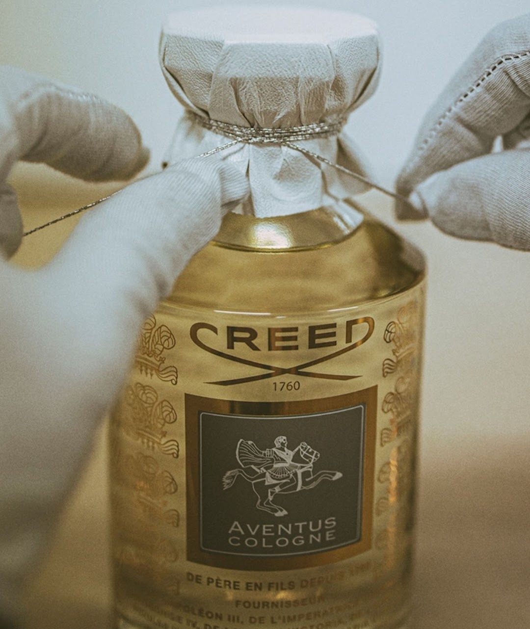 A close-up of a Creed Aventus Cologne bottle with a gold-toned body and white fabric-wrapped cap, being tied with a string by gloved hands. The front label shows the Creed logo and Aventus Cologne text.