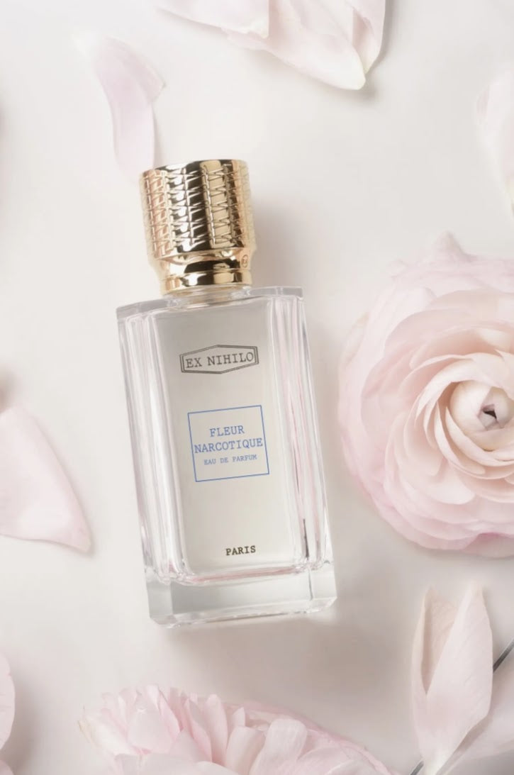 A clear glass perfume bottle with a textured gold cap, labeled 'EX. NINOLLO' and 'FLEUR NARCIOTIQUE', placed on a white background with soft pink rose petals.

