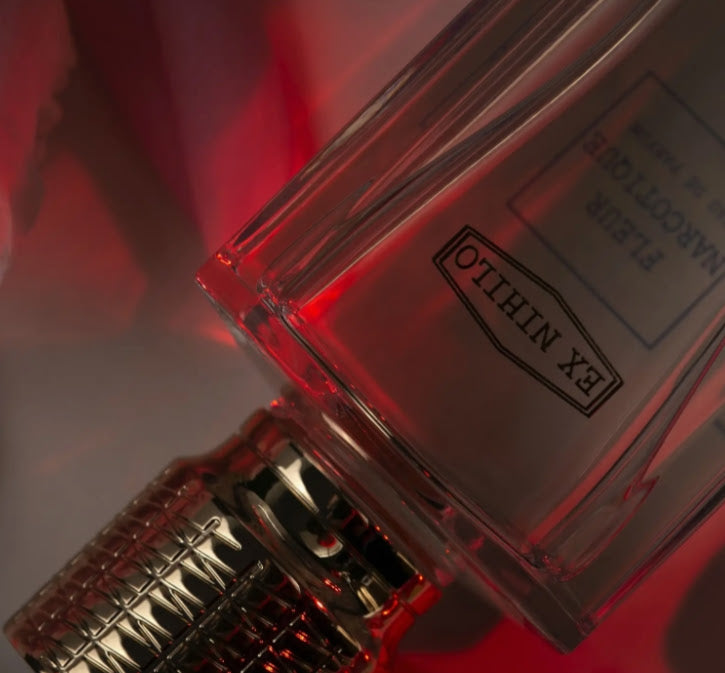 A close-up of a clear rectangular perfume bottle with a textured metallic cap, lit by red ambient light. The bottle shows the brand name 'EX.