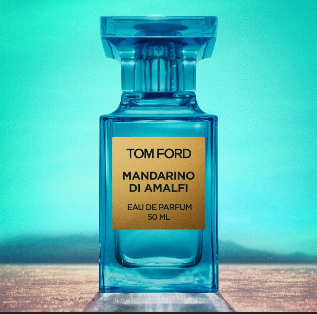 A Tom Ford Mandarin di Amalfi Eau de Parfum bottle, made of transparent blue glass with a gold label, set against a turquoise background.

